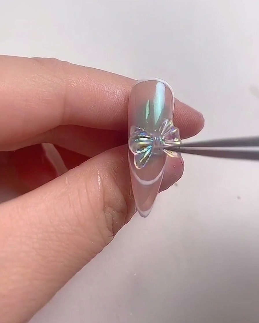 Unas pinzas de metal colocando cuidadosamente un brillante lazo de cristal iridiscente en el centro de una uña almendrada.
