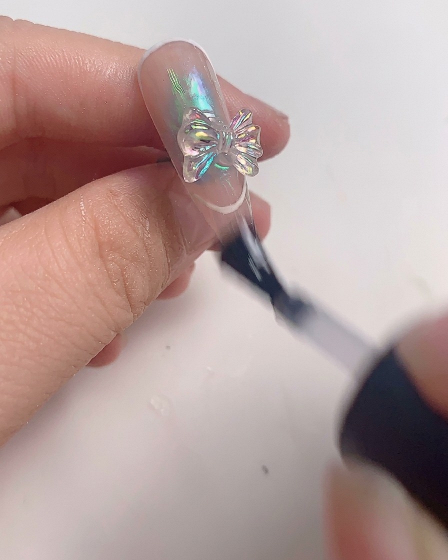 Un pincel aplicando una capa final de top coat transparente alrededor de un dije de lazo de cristal en una uña terminada.