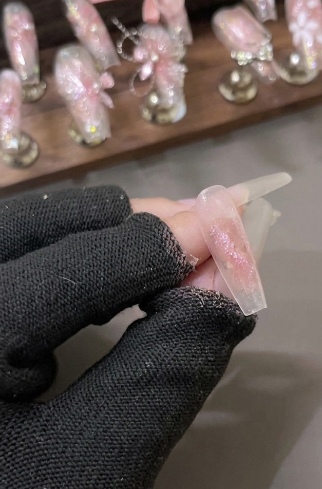 Una mano con guantes sosteniendo una uña transparente cubierta con una capa brillante de gel de purpurina de diamante triturado rosa.