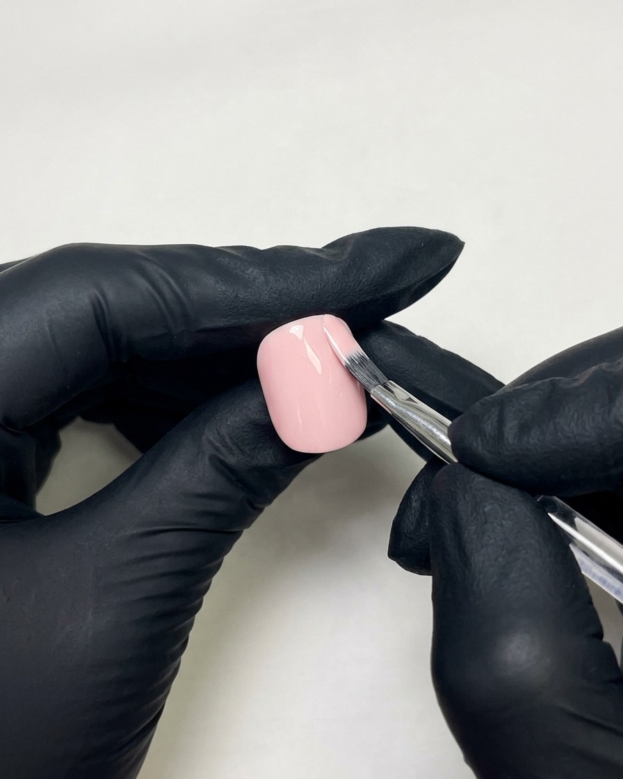 Manos con guantes usando un pincel plano para aplicar esmalte de gel rosa piel sobre una uña preparada.