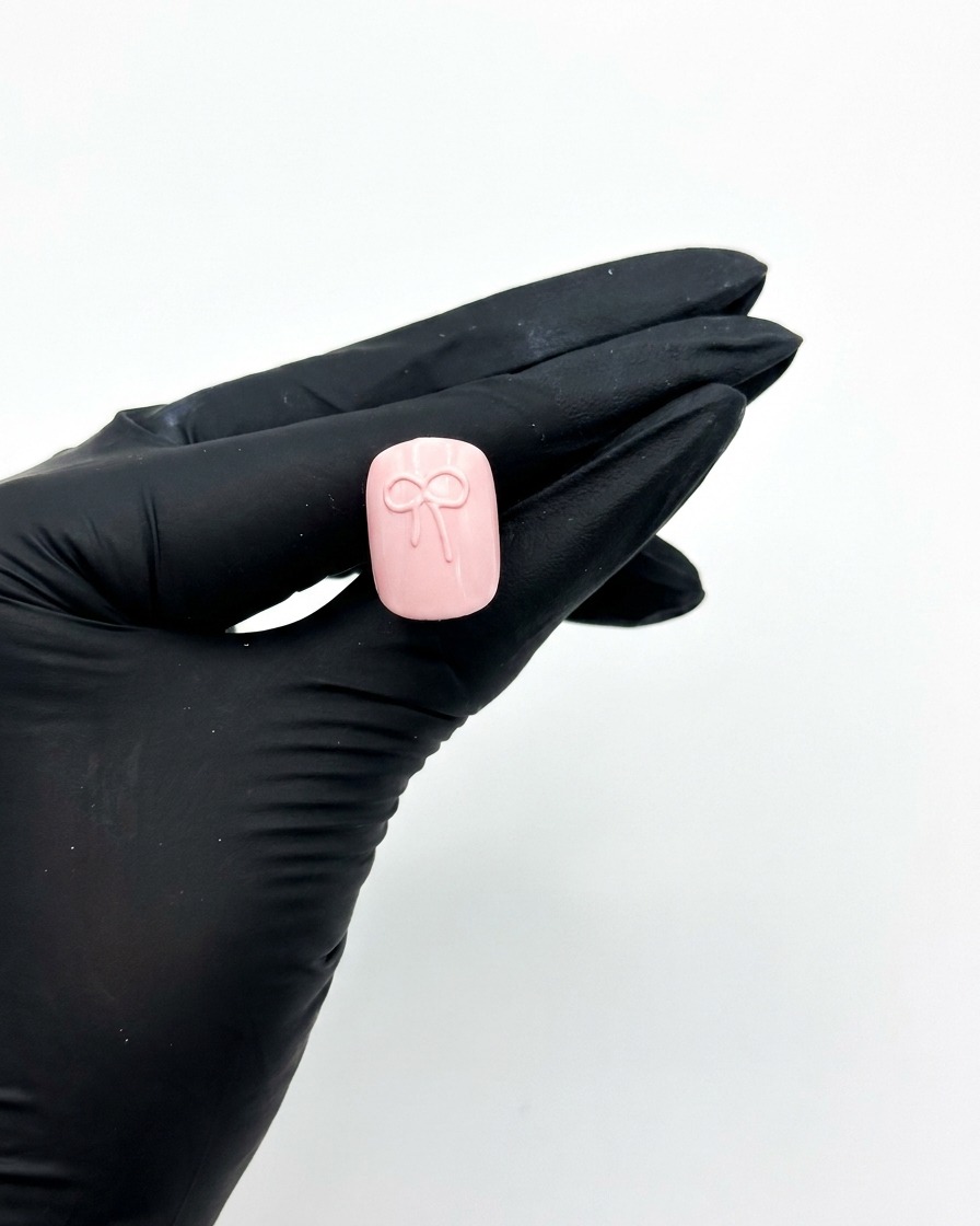 Mano con guantes sosteniendo una uña postiza rosa bajo una lámpara de uñas UV para curar el diseño del lazo.