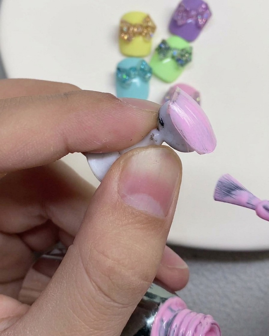 Un pincel aplicando esmalte en gel de tono rosa pastel suave sobre la superficie de una uña postiza en su soporte.