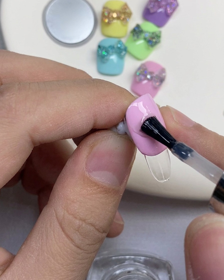 Un pincel negro aplicando gel de refuerzo transparente sobre una uña con esmalte rosa.