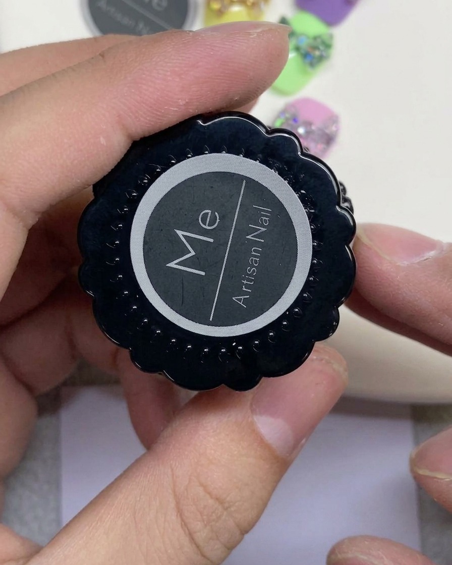 Una mano sosteniendo un tarro negro etiquetado como Artisan Nail con un logotipo circular blanco.