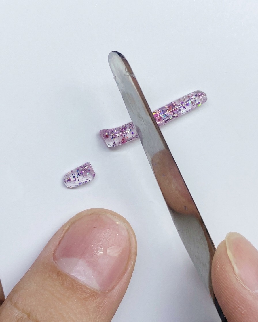 Una espátula de metal cortando una tira larga de gel PVC rosa brillante en segmentos más pequeños sobre una superficie blanca.