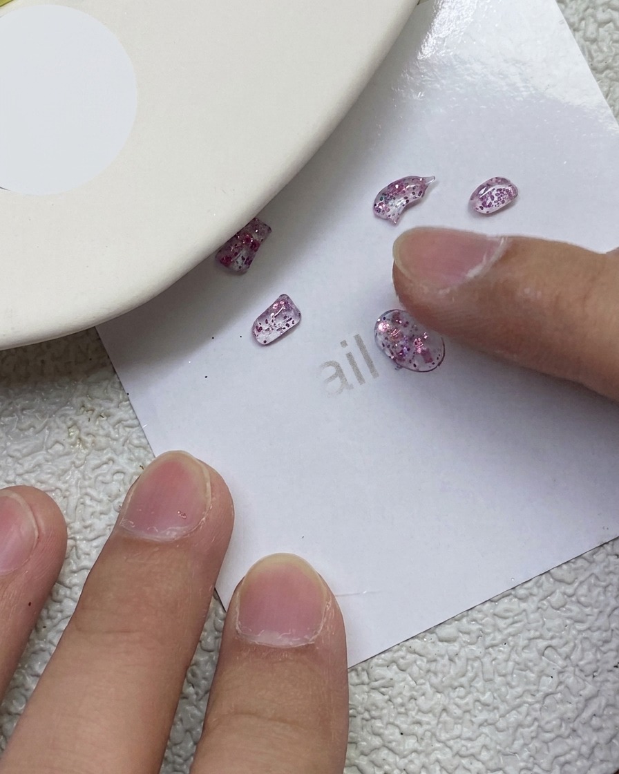 Un dedo presionando y esculpiendo pequeños trozos de gel PVC rosa brillante en forma de lazo sobre una hoja de papel blanca.