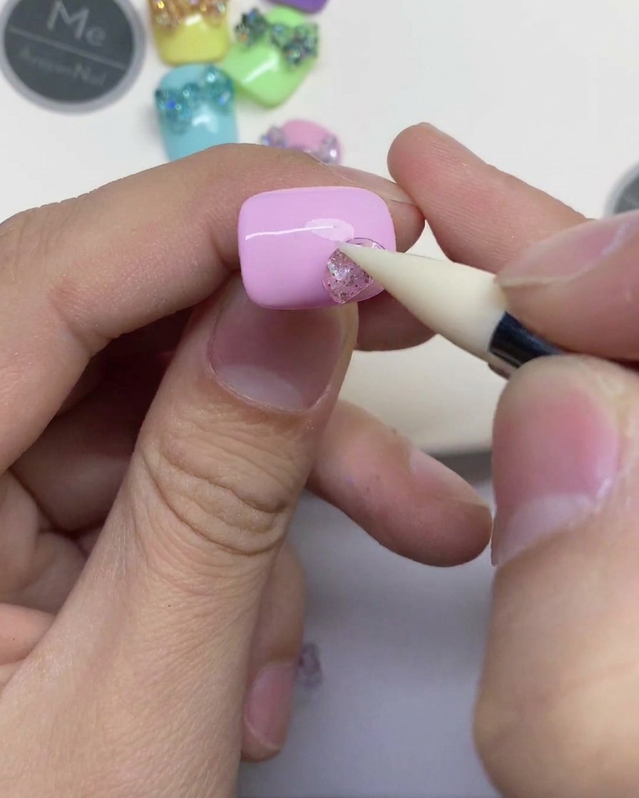 Primer plano de una mano utilizando una herramienta de silicona blanca para presionar una pequeña pieza de gel transparente con purpurina sobre una uña rosa claro.