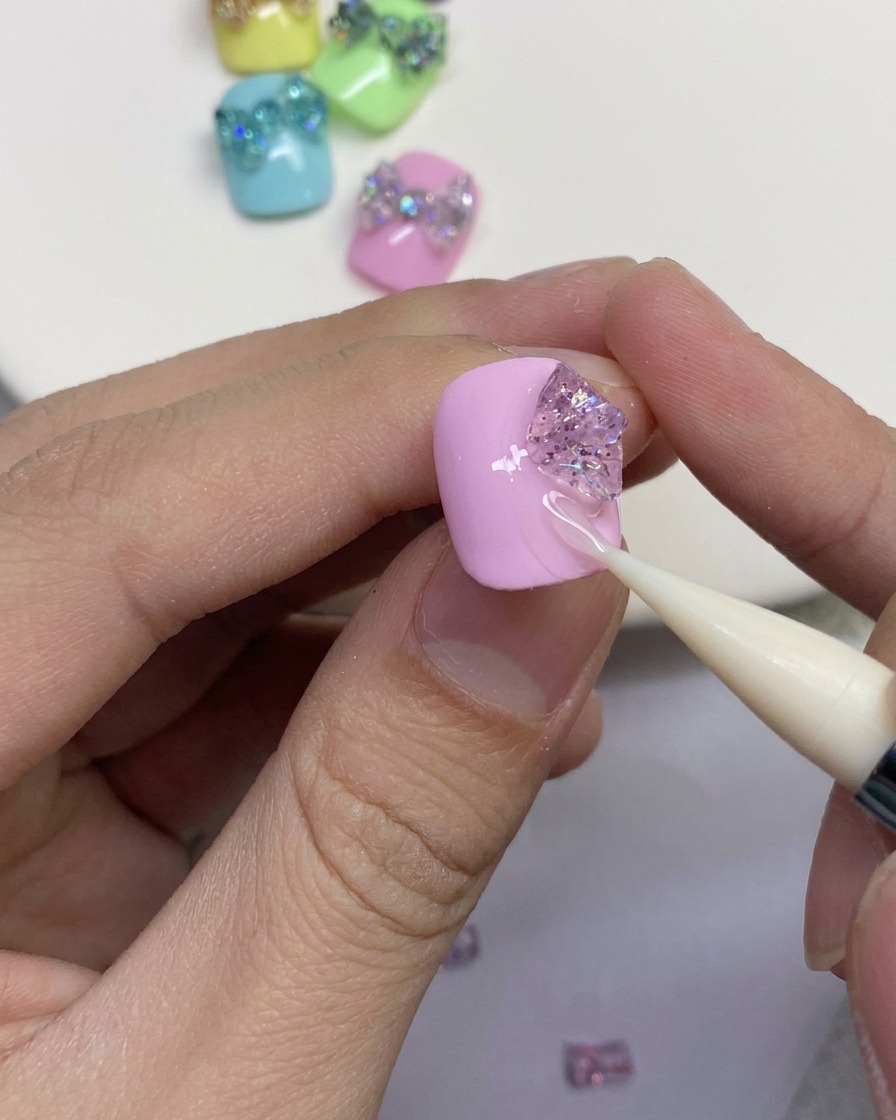 Primer plano que muestra una herramienta de silicona dando forma a piezas de gel con purpurina para formar los bucles exteriores de un lazo de mariposa sobre una uña rosa.