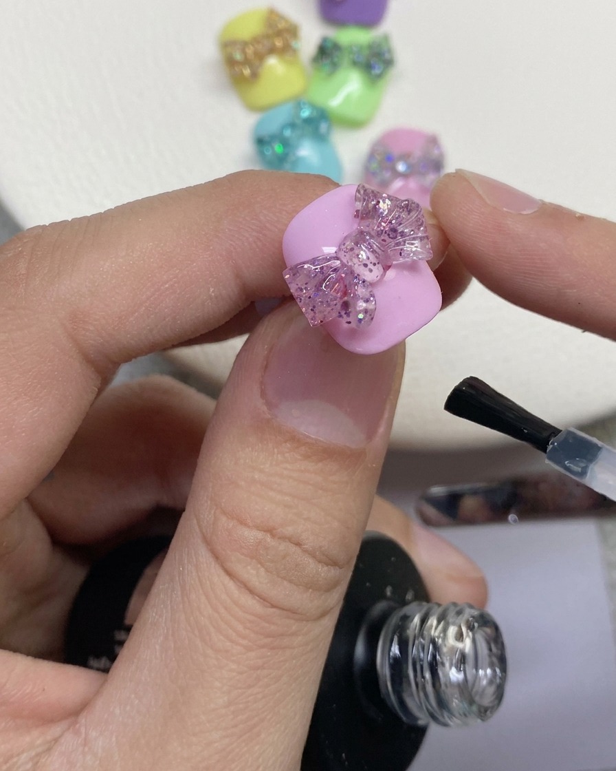 Primer plano de un pincel de uñas aplicando una capa de acabado transparente alrededor de un lazo brillante en 3D sobre una uña pintada de rosa.