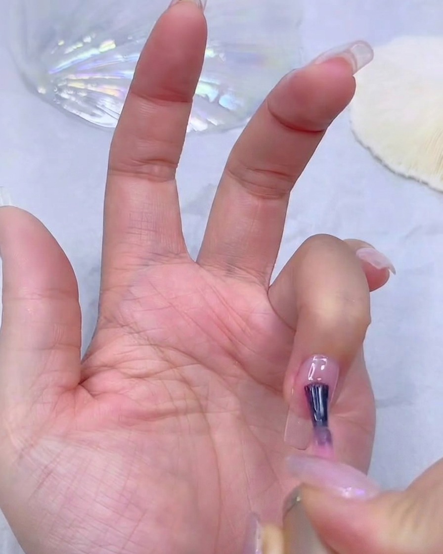 Mano de una técnica de uñas aplicando una capa base de gel rosa translúcido sobre una extensión de uña transparente.