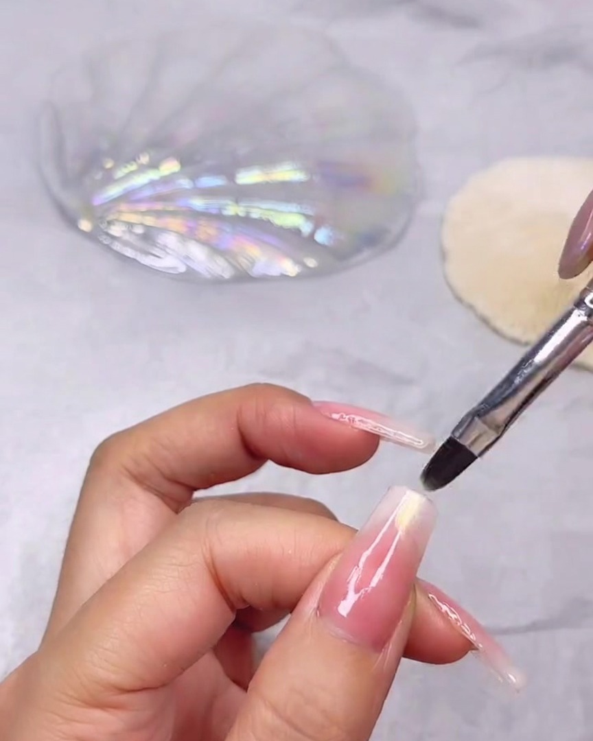 Un pincel fino de uñas aplicando un brillante gel transparente con textura de concha en la punta de una uña larga con degradado rosa y blanco.