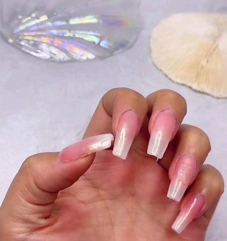 Primer plano de una mano mostrando uñas largas y brillantes con un degradado rosa y blanco lechoso, destacando un gran brillante de corazón cerca de la cutícula del pulgar.