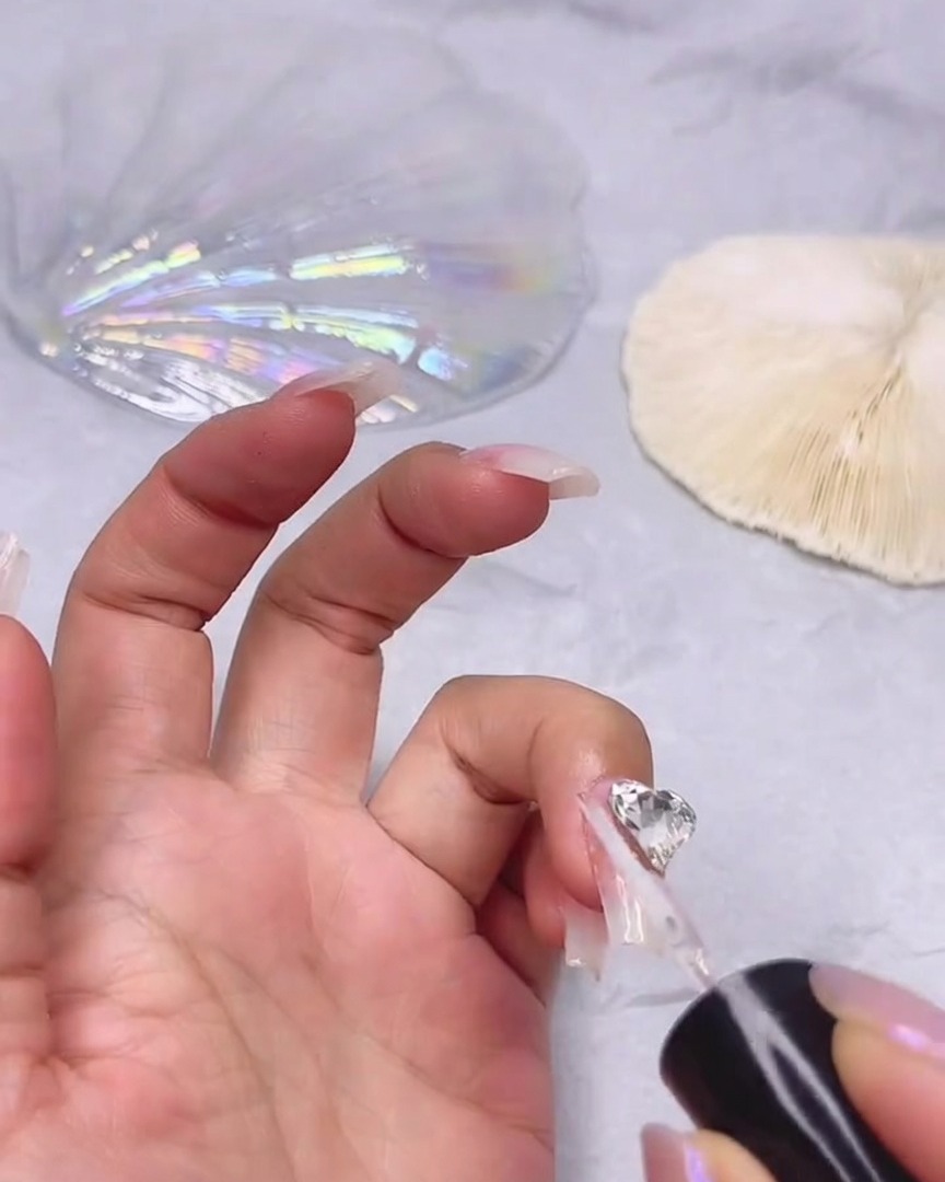 Un pincel fino aplicando gel sellador transparente alrededor de los bordes de una gran pedrería transparente en forma de corazón pegada a una uña.