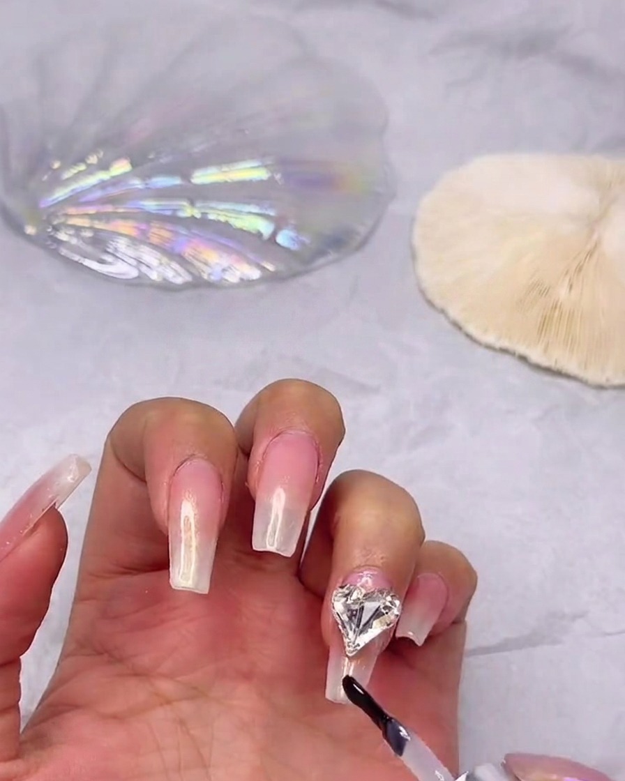Un pincel de esmalte en gel aplicando una capa de top coat brillante sobre una uña degradada, trabajando cuidadosamente alrededor de una pedrería de corazón fijada.