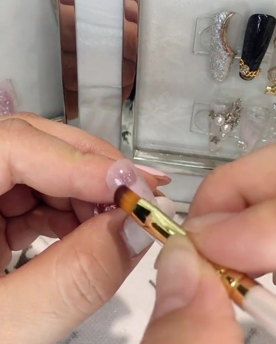 Una mano sosteniendo un pincel de uñas aplicando gel rosa en la mitad superior de una uña en forma de almendra.