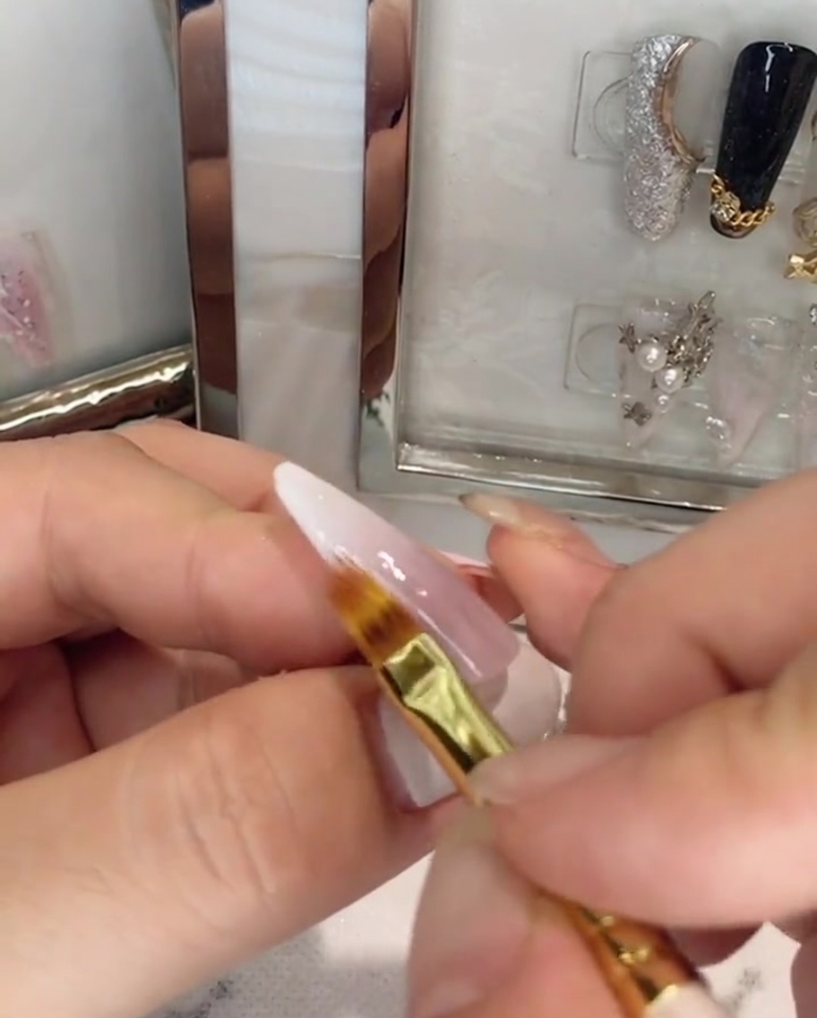 Un pincel aplicando capa superior transparente sobre una uña ombré rosa y blanca.