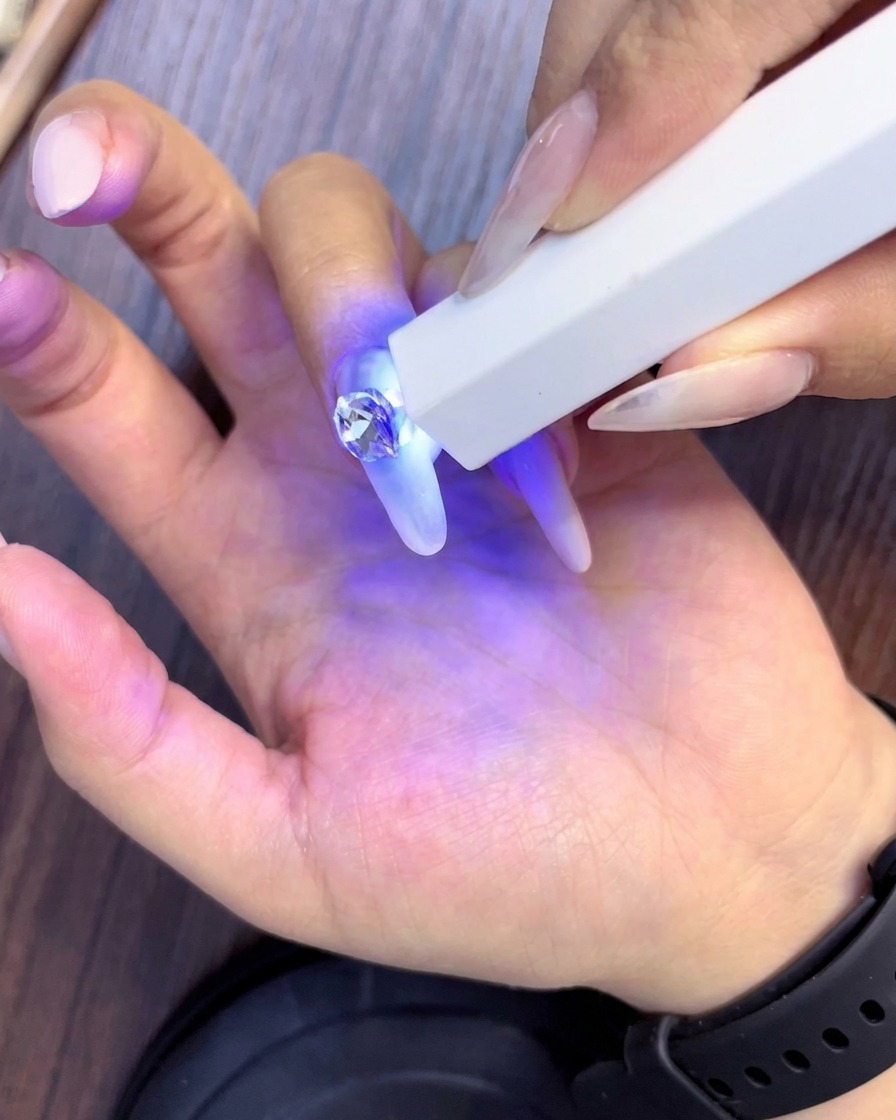 Una lámpara UV de mano curando la pedrería recién colocada que rodea al cristal central principal en una uña con forma de almendra.