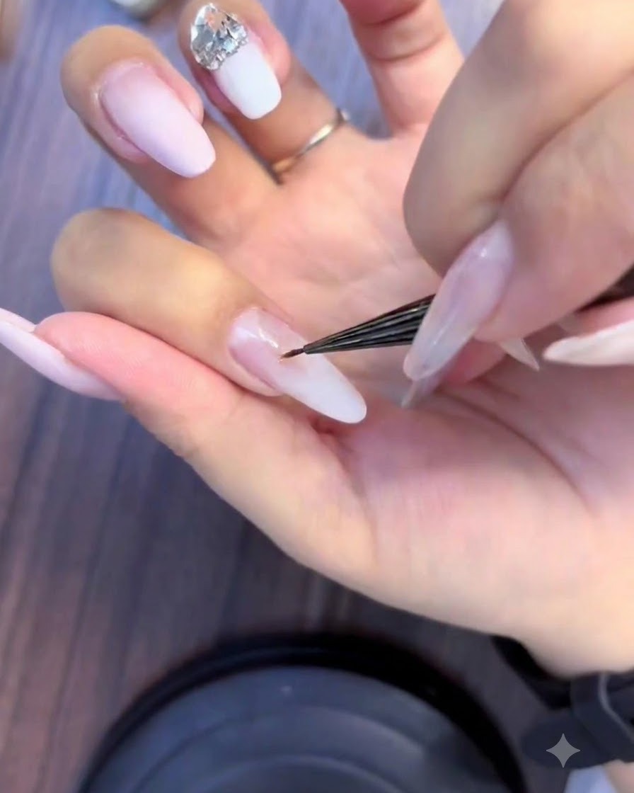 Un primer plano de una técnica de uñas usando un pincel fino para aplicar pedrería pequeña en un grupo de cristales en una uña ombré rosa y blanca.