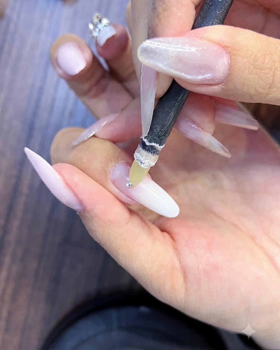 Una herramienta de lápiz de recogida de cera sosteniendo un diminuto diamante de imitación plateado sobre una uña preparada con degradado rosa y blanco.