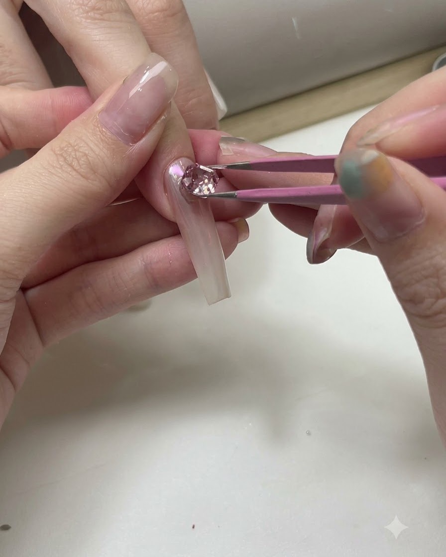 Pinzas colocando cristales rosas y transparentes sobre un tip de uña de práctica transparente.