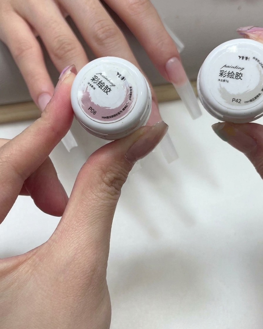 Manos sosteniendo dos pequeños frascos de gel de color rosa y blanco, con uñas esculpidas al fondo.