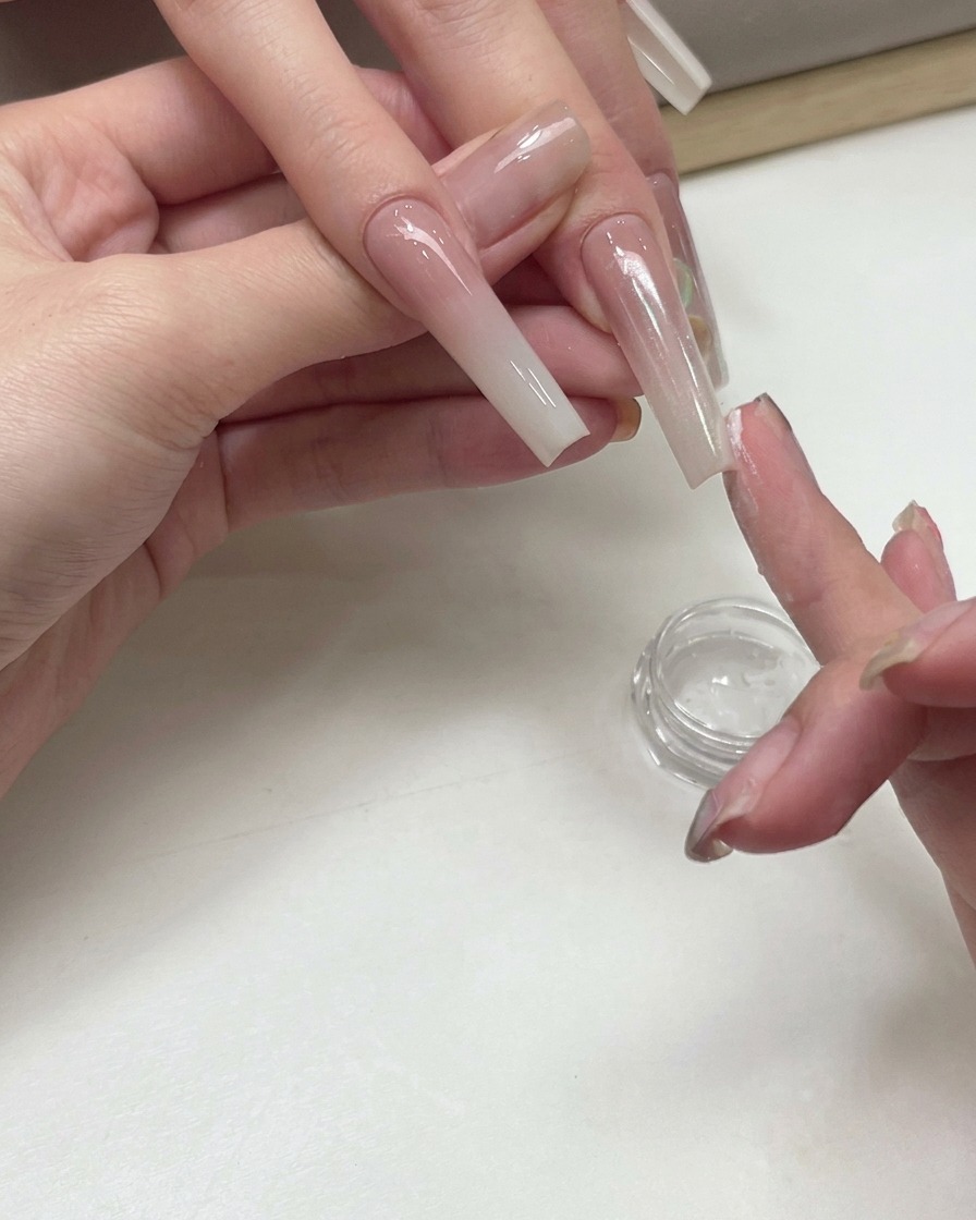 Manicurista usando la yema del dedo para aplicar polvo de pigmento brillante tono melocotón sobre una uña degradada.