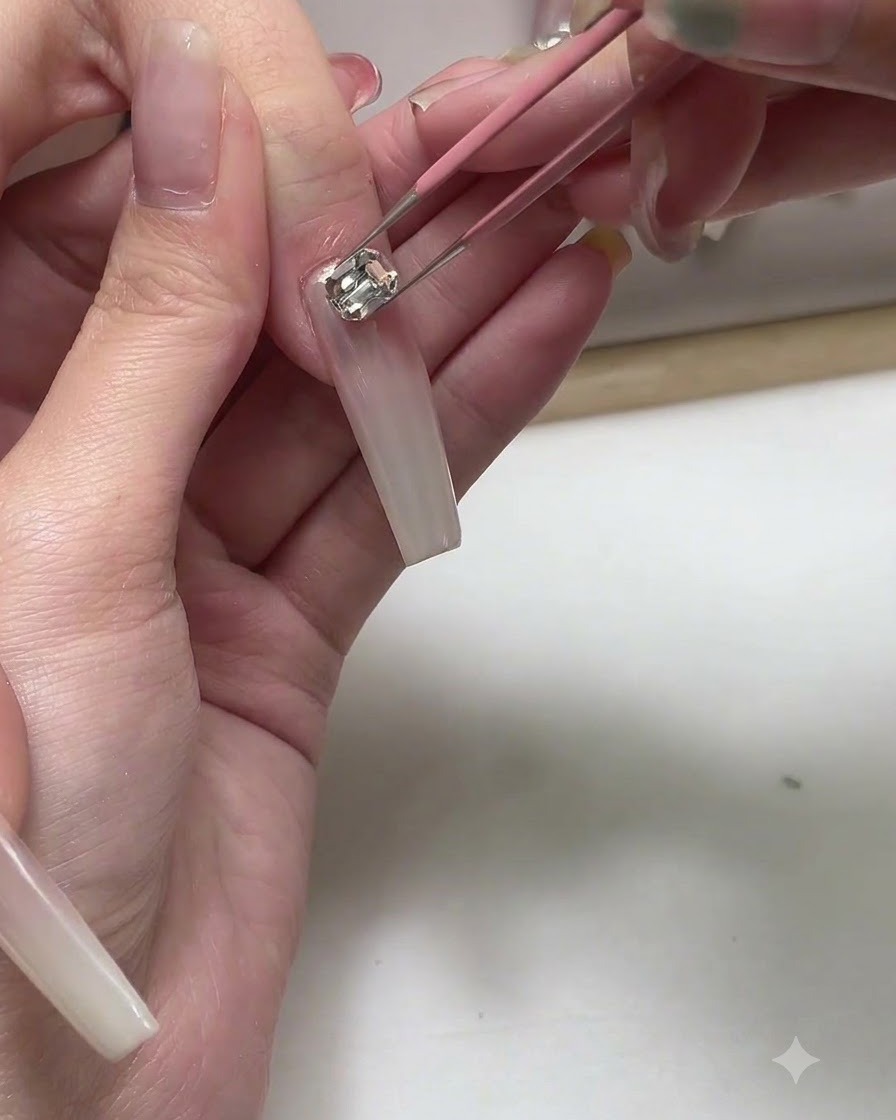 Pinzas colocando un pequeño dije metálico sobre una uña larga de gel en tono rosa nude.