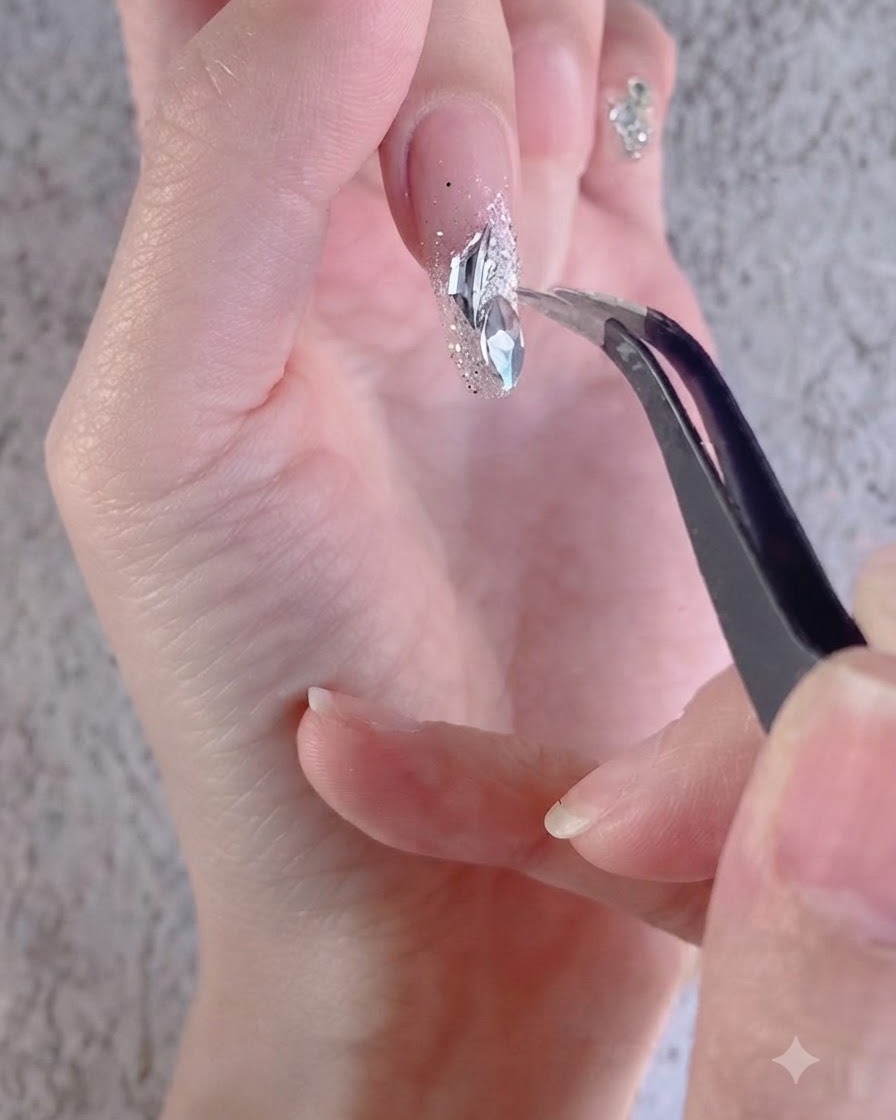 Una mano utilizando unas pinzas para ordenar pedrería blanca irregular sobre una uña decorada.