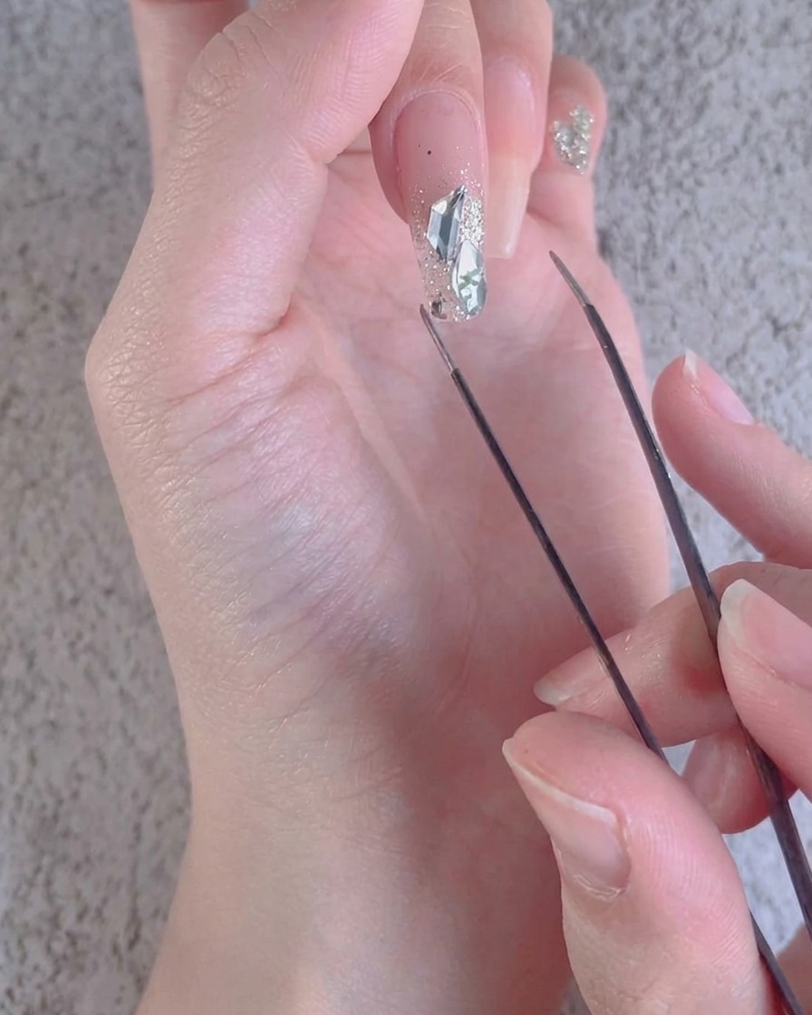 Un primer plano de unas pinzas ajustando pequeños detalles de diamante triturado alrededor de piedras más grandes en una uña.