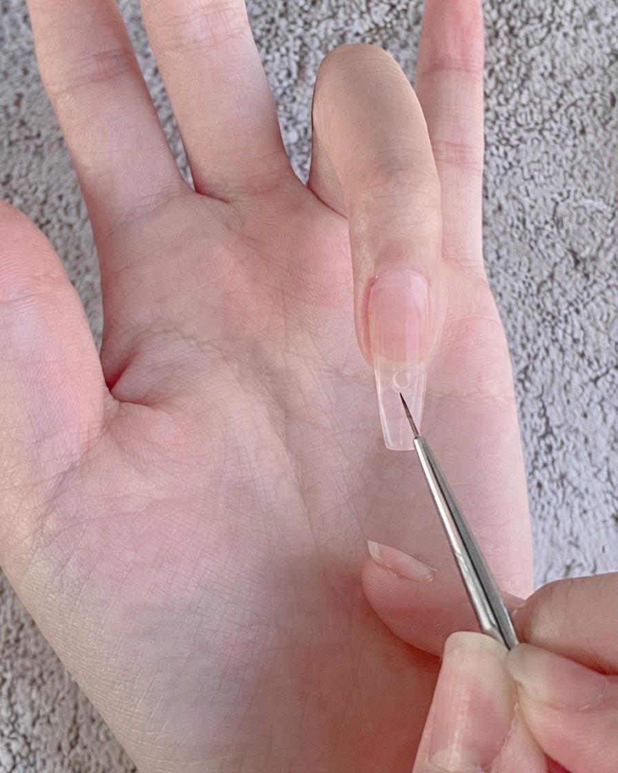 Un pincel delineador fino de nail art colocando una pequeña gota transparente de gel para pedrería sobre una uña recubierta de gel nude.