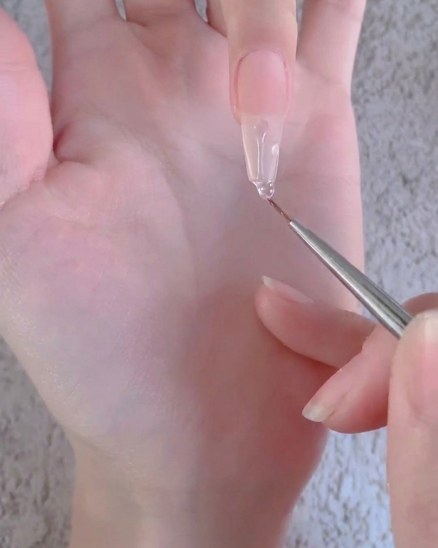 Un pincel delineador tirando de una gota de gel transparente hacia abajo a lo largo de una extensión de uña artificial larga.