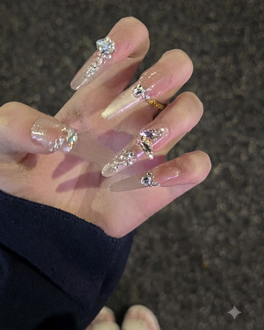Primer plano de una mano con uñas largas nude adornadas con pedrería, ligeramente curvadas para mostrar el perfil.