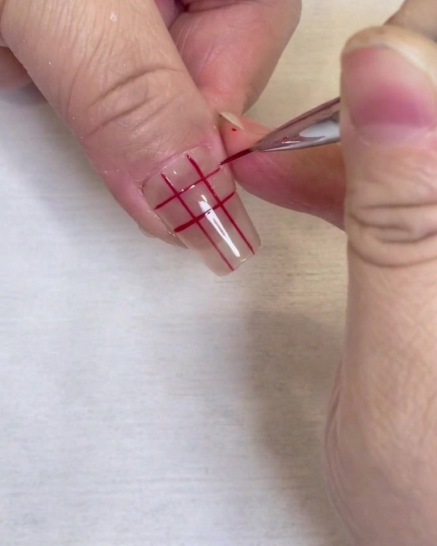 Un primer plano de un patrón de cuadrícula roja completado en una uña transparente, dibujado con un pincel delineador fino.