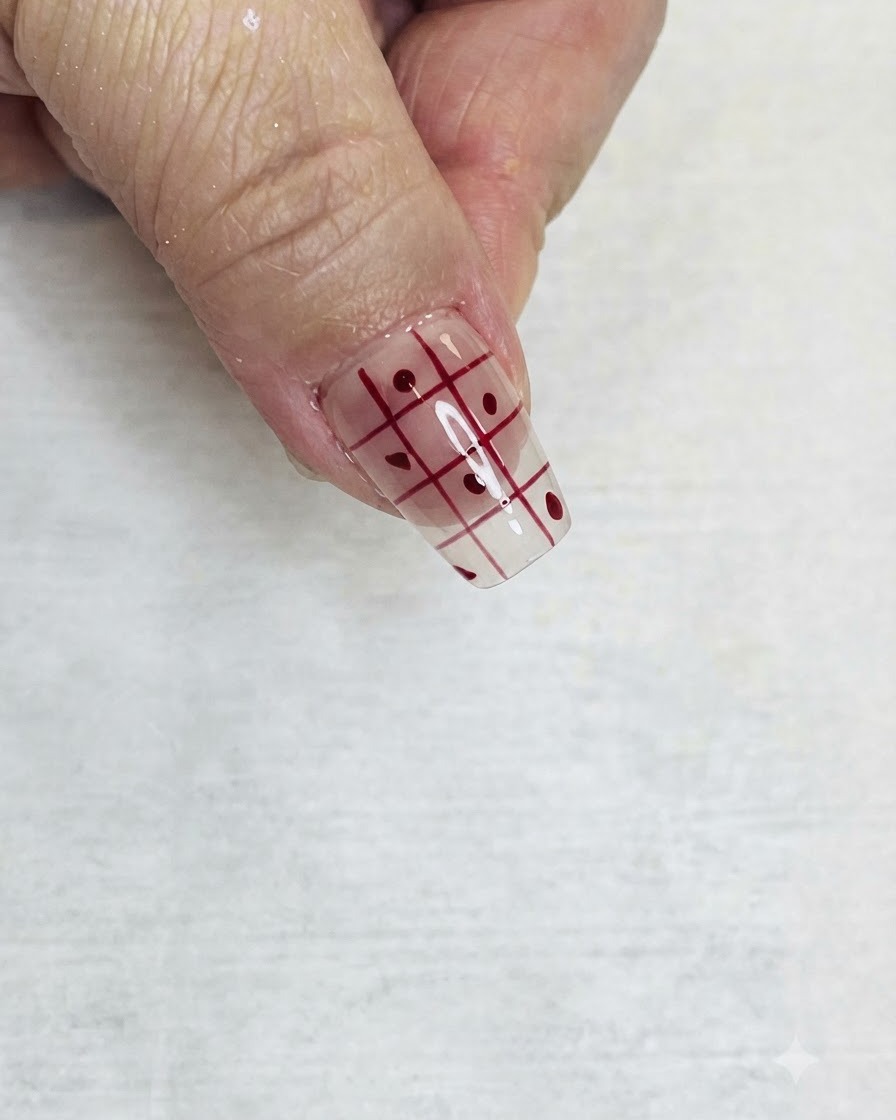 Un pincel delineador fino rellenando cuadrados alternados de un patrón de cuadrícula roja en una uña transparente con esmalte rojo.