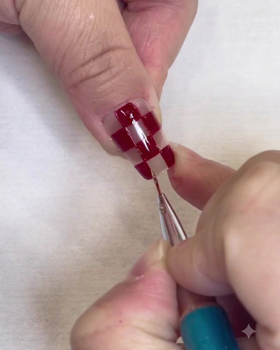 Un pincel de detalles finos pintando bordes delgados de brillo plateado a lo largo de los bordes de los cuadrados rojos en una uña transparente.