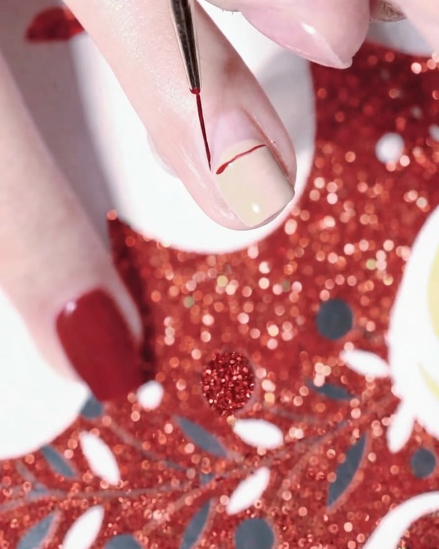 Primer plano de una mano usando un pincel delineador fino para dibujar una línea curva roja para el gorro de Papá Noel sobre una uña pintada de color nude.