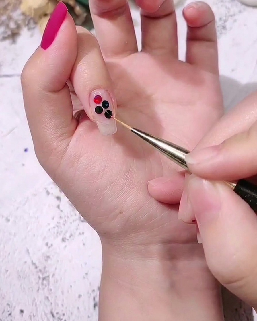 Un primer plano de una mano colocando lentejuelas de puntos de colores medianas junto a otras más grandes en una uña usando un pincel de detalle fino.