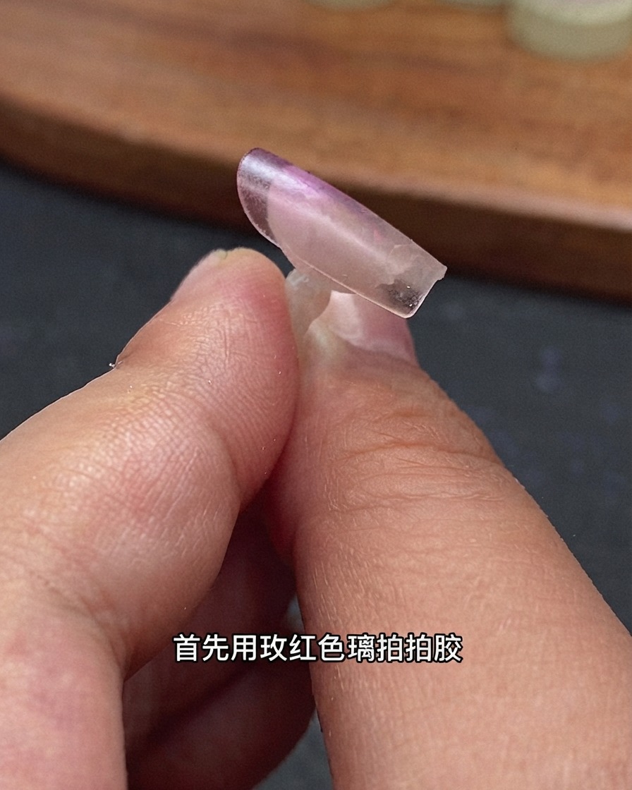 Primer plano de una punta de extensión de uña transparente sostenida por los dedos con gel efecto vidrio rojo rosa aplicado en el borde posterior.