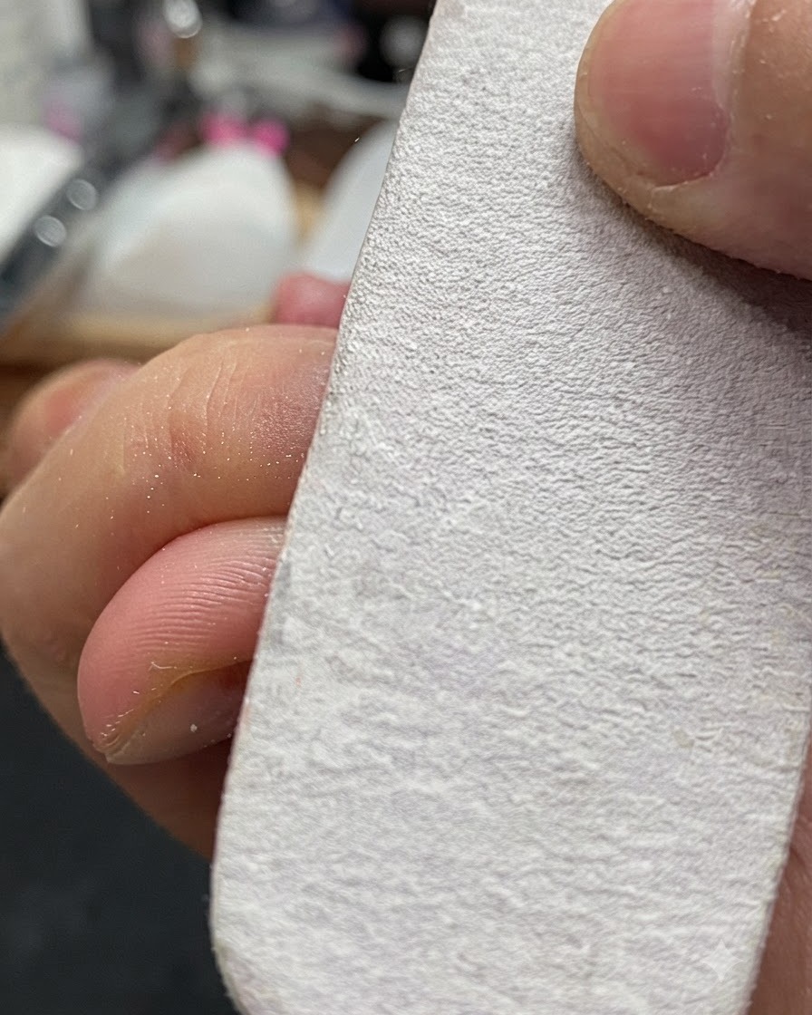 Una mano sosteniendo un bloque pulidor de uñas blanco limando ligeramente la superficie de una uña natural.