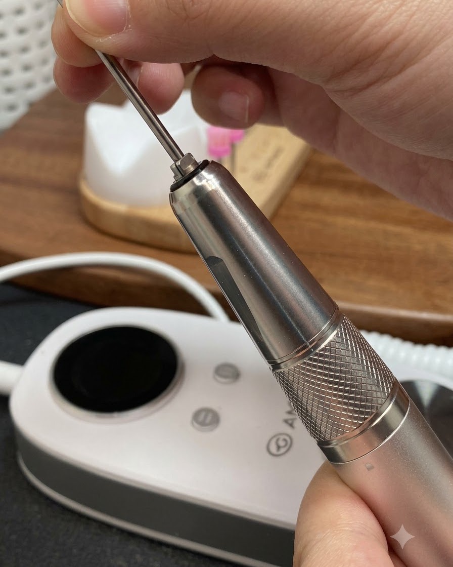 Manos insertando una broca metálica cónica larga en la pieza de mano de una lima de manicura eléctrica.