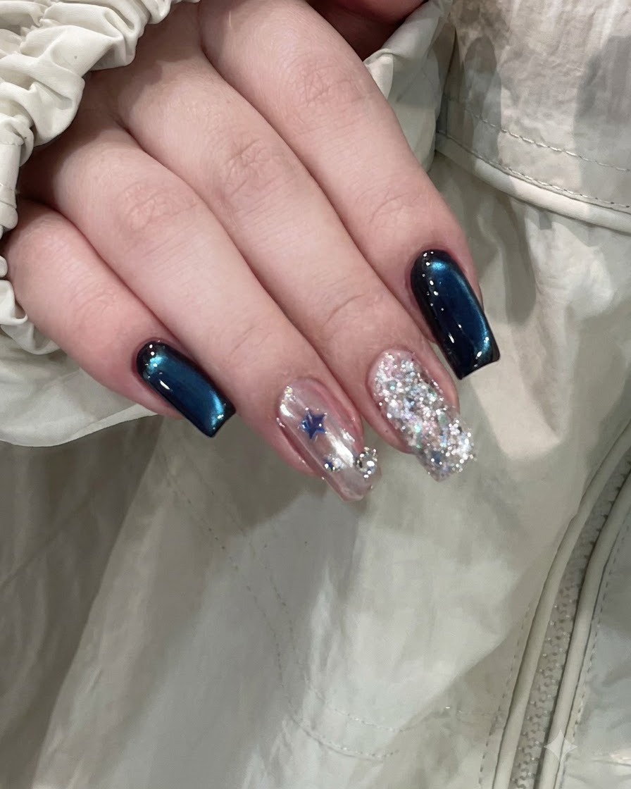 Una mano terminada que muestra uñas de color azul intenso en ojo de gato junto a una uña acentuada con estrellas y otra completamente cubierta de purpurina.