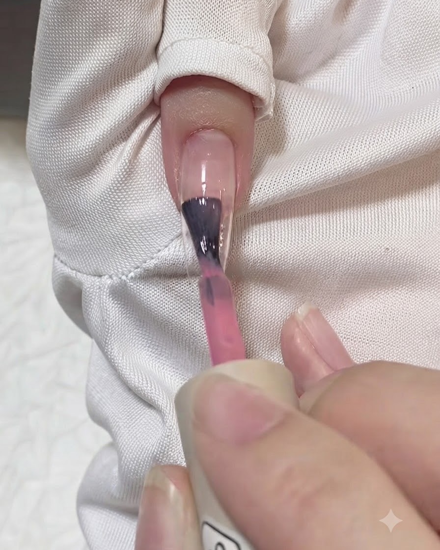 Un pincel aplicando una capa de gel rosa translúcido en el centro de una extensión de uña transparente.