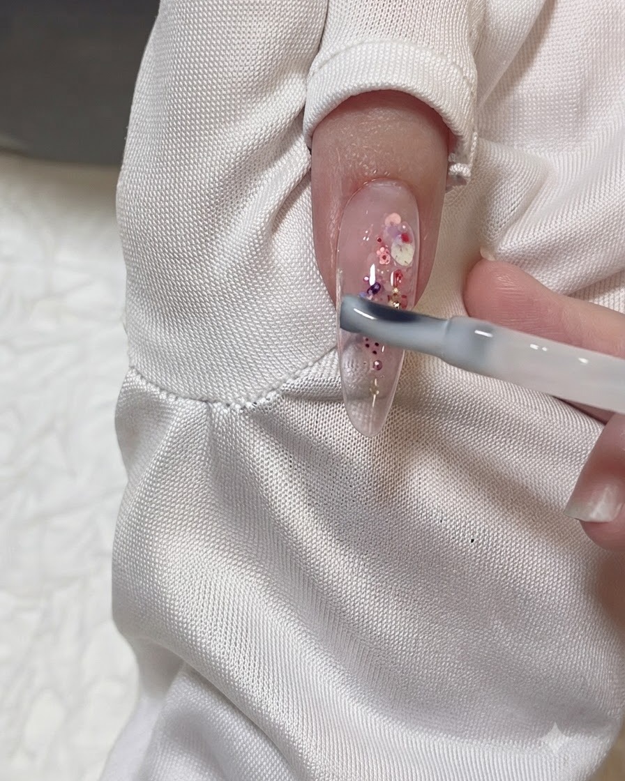 Un pincel aplicando gel lechoso sobre una uña almendrada con purpurina rosa y morada.