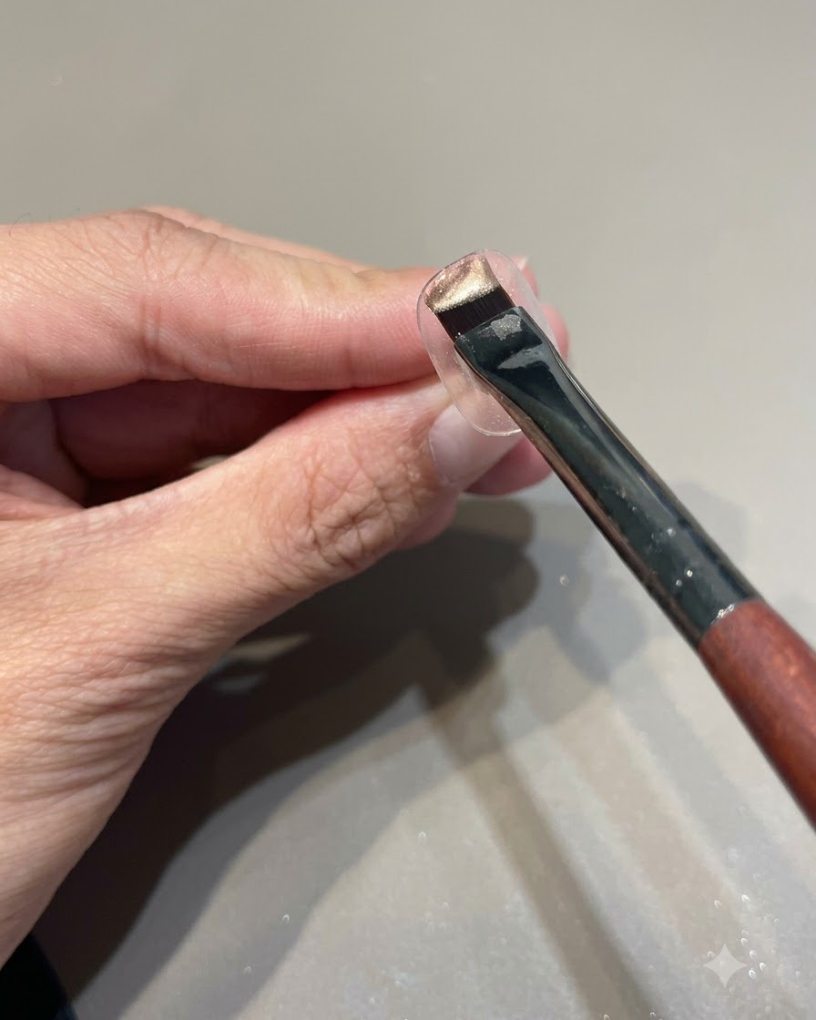 Un pincel cargado con esmalte de gel ojo de gato rosa dorado sostenido sobre un soporte transparente para exhibir uñas.