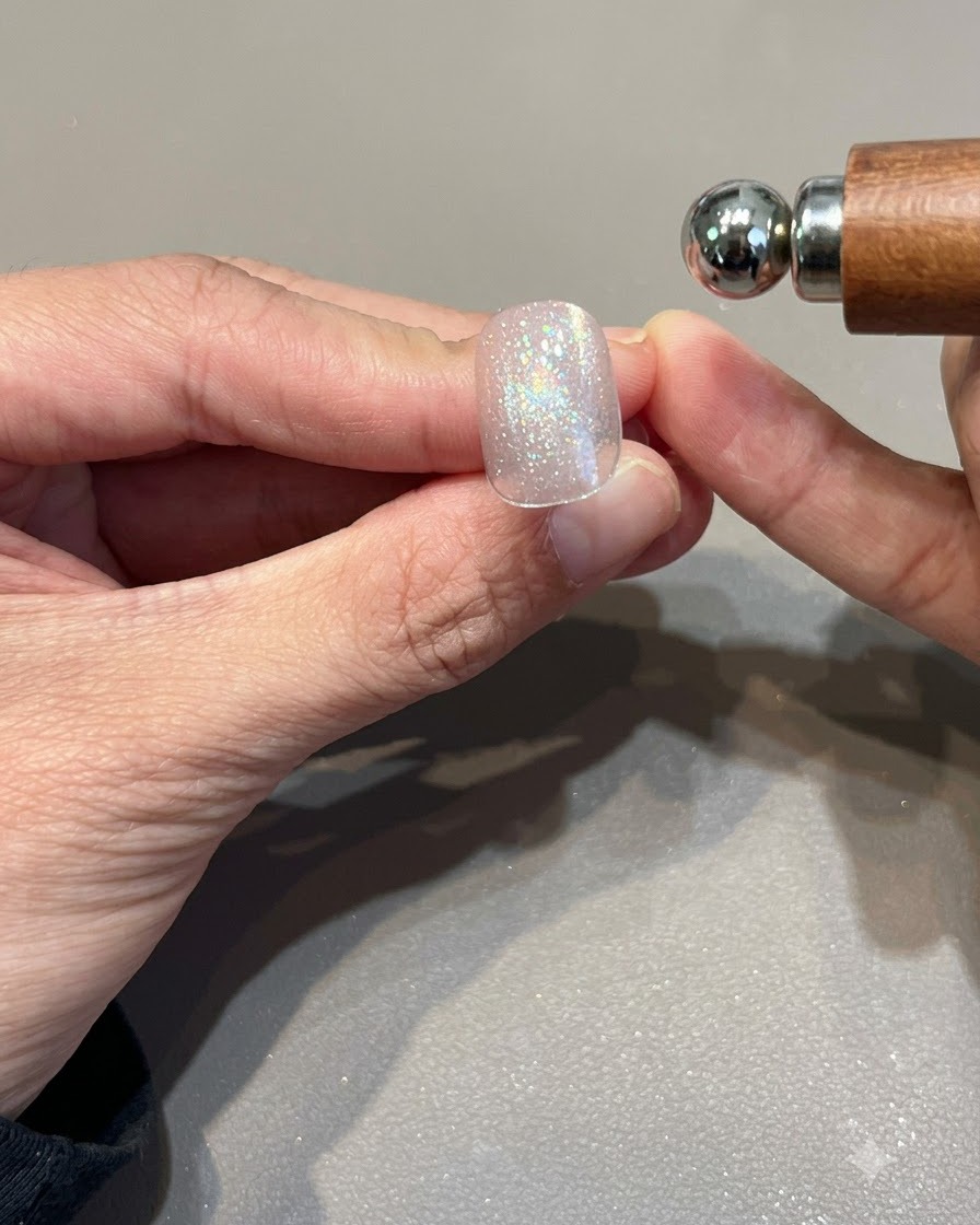 Un imán esférico sostenido cerca de una uña de gel ojo de gato rosa dorado brillante para dar forma a la banda de luz.