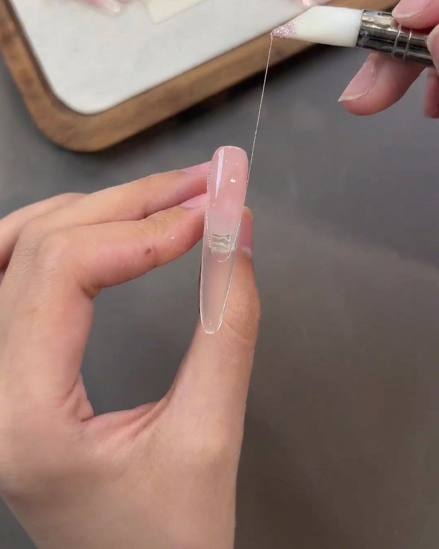 Un pincel trazando una fina hebra de gel rosa con purpurina a lo largo de la superficie de una uña en tono nude.