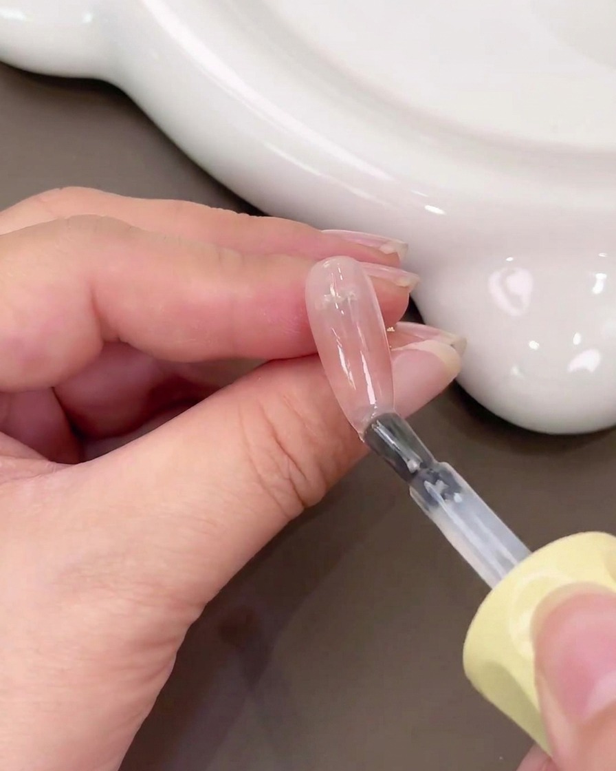 Un pincel aplicando gel base transparente sobre una uña con un degradado de blush rosa.