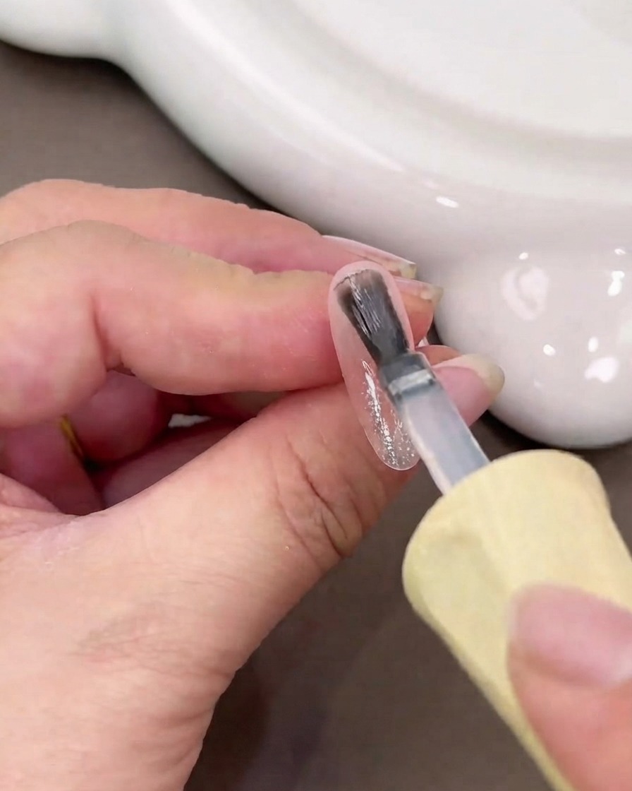 Un pincel de uñas aplicando una capa húmeda de gel base transparente sobre una superficie de uña brillante.