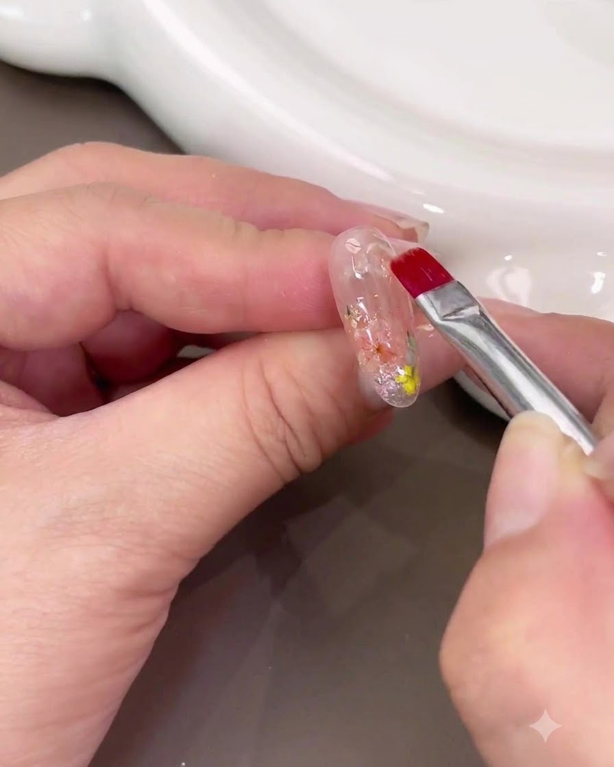Un pincel de uñas aplicando una capa gruesa de gel de construcción transparente sobre el nail art de flores secas.