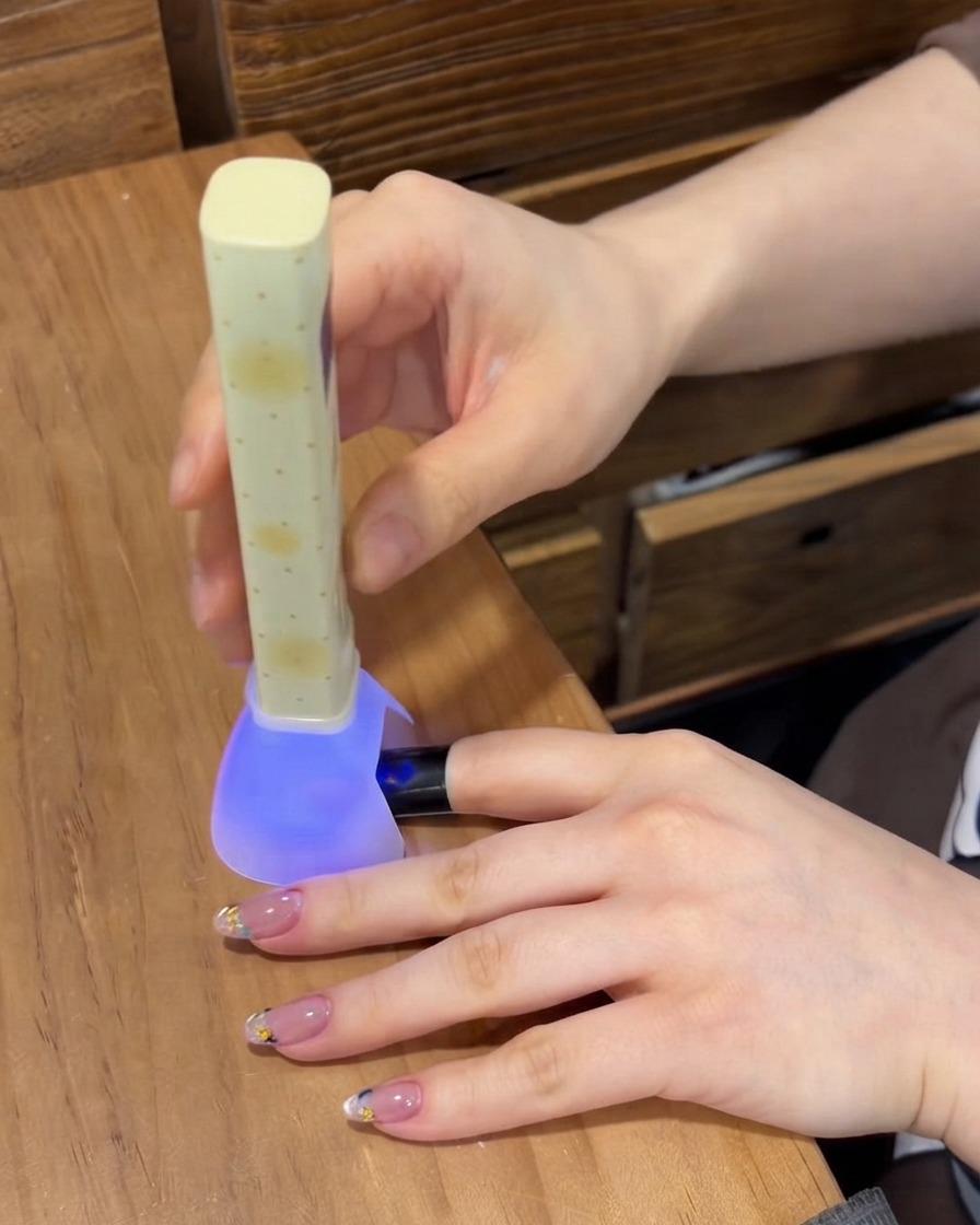 Una lámpara de uñas portátil UV/LED morada curando el gel en una uña mientras los otros dedos descansan sobre la mesa.